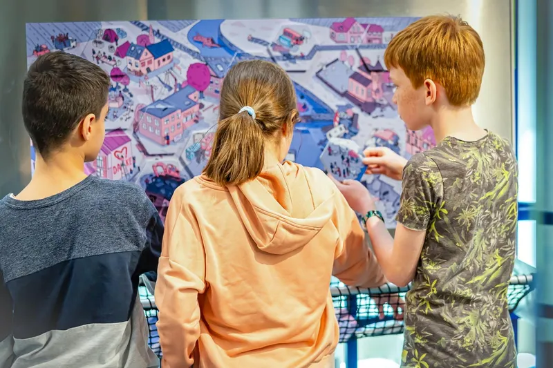 Drei Kinder erkunden ein illustriertes Puzzle mit einer Dorfszene.