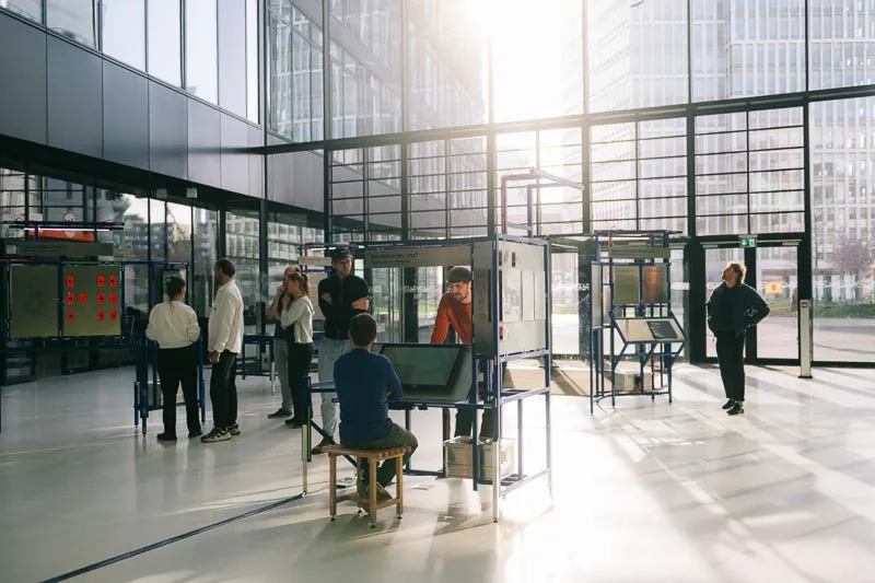 A group of visitors gathers around the exhibition stations in a sunlit, glass-walled atrium.