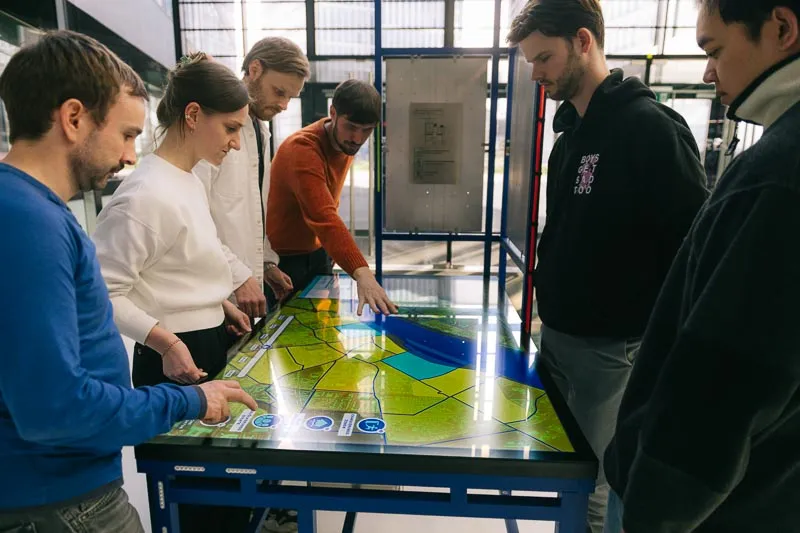 Six people stand around an interactive table showing a map in a bright exhibition hall.