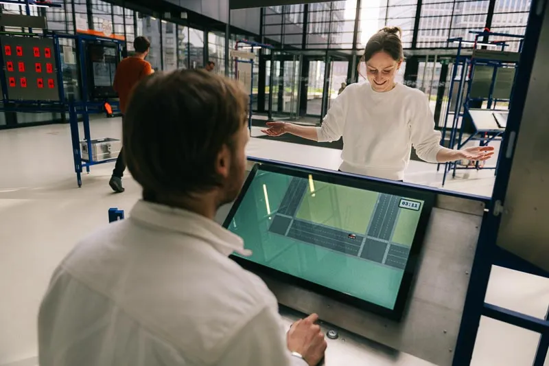 Two people play a game on opposite sides of the interactive station. A man operates a touchscreen with a street map while a smiling woman stands opposite.