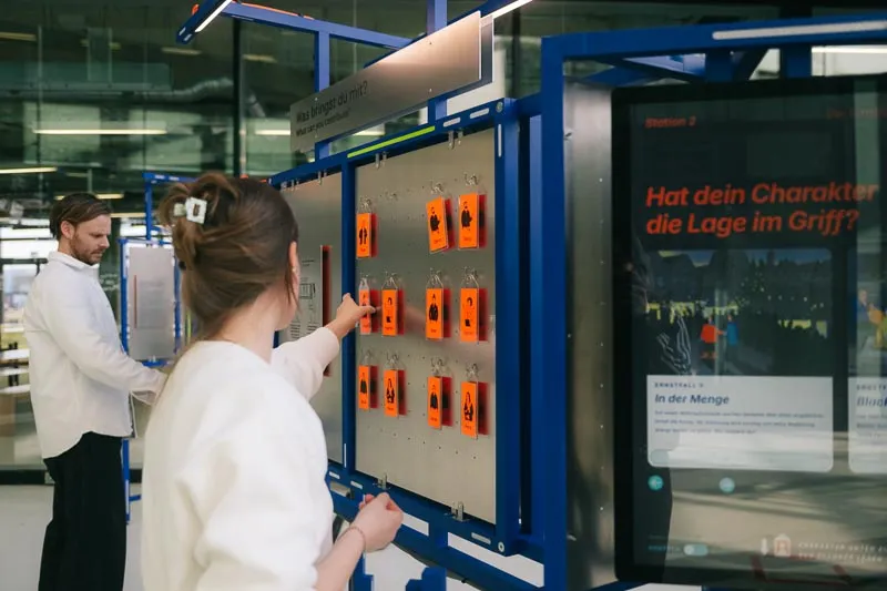 Two people at the station. One hangs orange tokens on a pegboard beside a touchscreen.