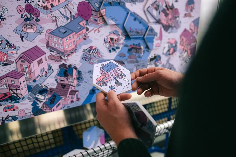 Hands hold a hexagonal illustrated puzzle tile over a drawn map of a flooded town.